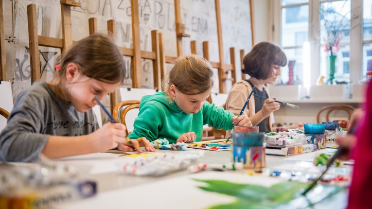 Dzieci siedzące przy stole podczas zajęć plastycznych, skupione na malowaniu i rysowaniu w pracowni artystycznej.