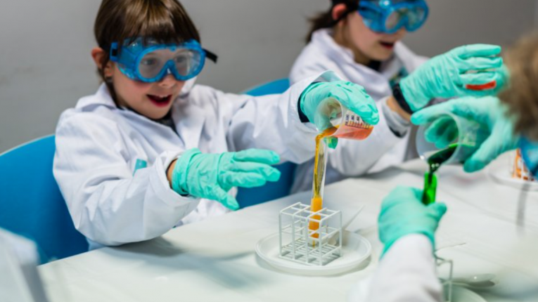 Dwoje dzieci ubranych w stroje chemików wykonuje doświadczenie chemiczne (przelewają płyny z naczyń do naczyń).