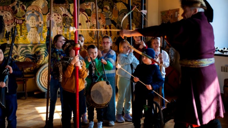 Dzieci stoją w półkolu, każde trzyma historyczną broń lub rekwizyt. Instruktor w stroju z epoki napina łuk, pokazując, jak strzelać. W tle kolorowa tkanina z dawnymi wzorami.