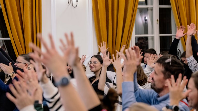 Uczestnicy i uczestniczki wydarzenia siedzą w sali i unoszą ręce. W tle widoczne są duże okna zasłonięte żółtymi kotarami.