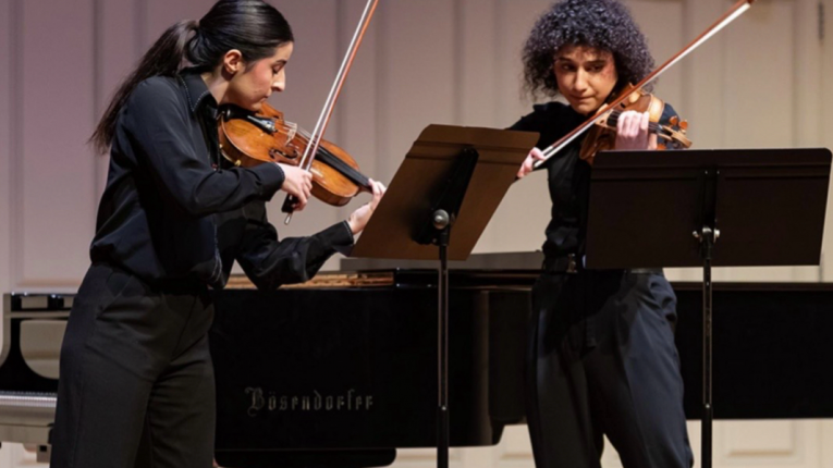 Dwie skrzypaczki w czarnych strojach grają w duecie na scenie koncertowej, stojąc przy pulpitach z nutami; w tle widoczny fortepian koncertowy.