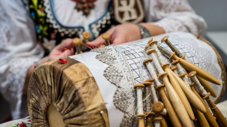 stelaż do koronki klockowej, a na nim koronka w trakcie tworzenia. Widać dłonie autorki odzianej w ludowy strój.
