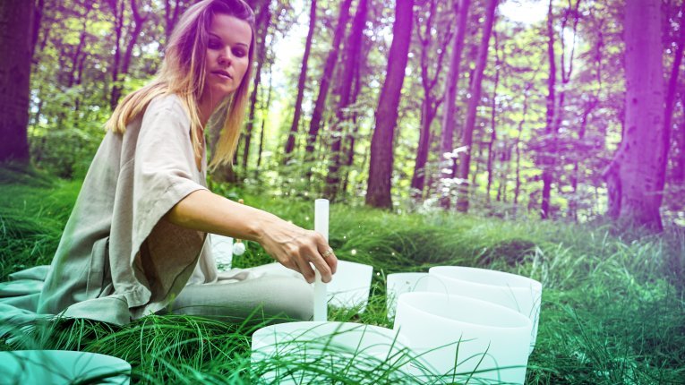 Kobieta siedząca na trawie w lesie, wykonująca kąpiel w dźwiękach przy użyciu białych mis kryształowych.