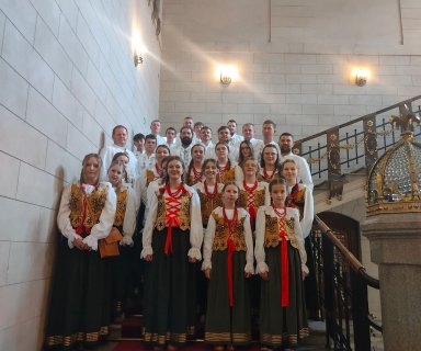 Zespół góralski w strojach tradycyjnych, stoi na schodach wewnętrznych w jasnym, przestronnym budynku.