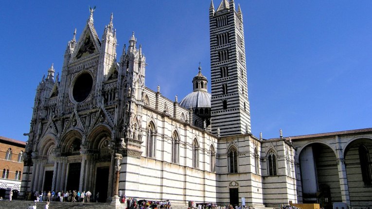 Fasada katedry w Sienie z biało-czarnymi pasami, wysoka dzwonnica i kopuła widoczne na tle bezchmurnego, niebieskiego nieba; na placu przed katedrą spacerują turyści.