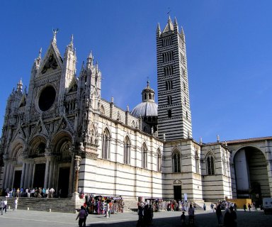 Fasada katedry w Sienie z biało-czarnymi pasami, wysoka dzwonnica i kopuła widoczne na tle bezchmurnego, niebieskiego nieba; na placu przed katedrą spacerują turyści.