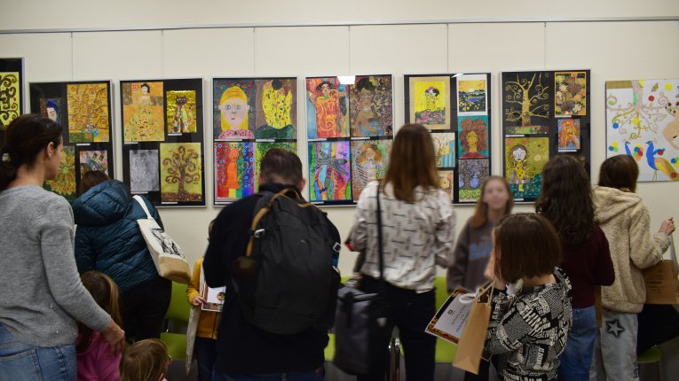 Ludzie widoczni tyłem stojący przed powieszonymi na ścianie pracami plastycznymi umieszczonymi w antyramy. Prace są kolorowe i różnorodne.