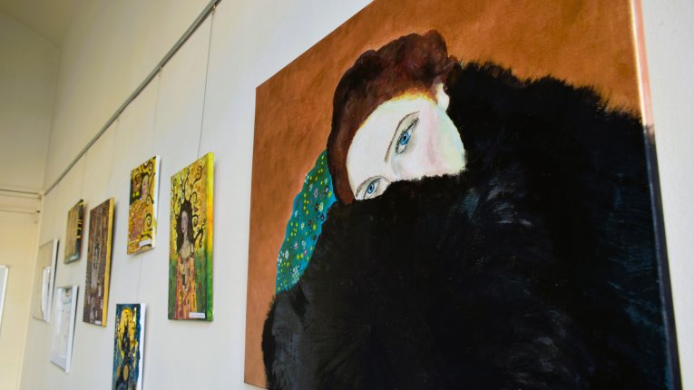 Obraz rudo-brązowowłosej, niebieskookiej kobiety o jasnej cerze, częściowo ukryta za czarnym futrem, które ją otula. Zza niej widzimy rude tło oraz fragment niebieskozielonej płaszczyzny pokrytej drobnymi obiektami - kwiatkami, ornamentami.