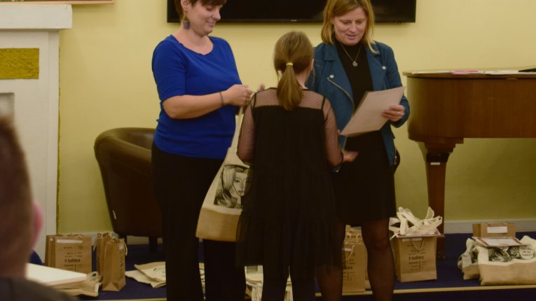 Widzimy organizatorki konkursu, przekazujące jednej z laureatek nagrodę oraz dyplom. W tle przyjemny obrazek łąki, ekran oraz fortepian. Klimat miejsca jest ciepły.