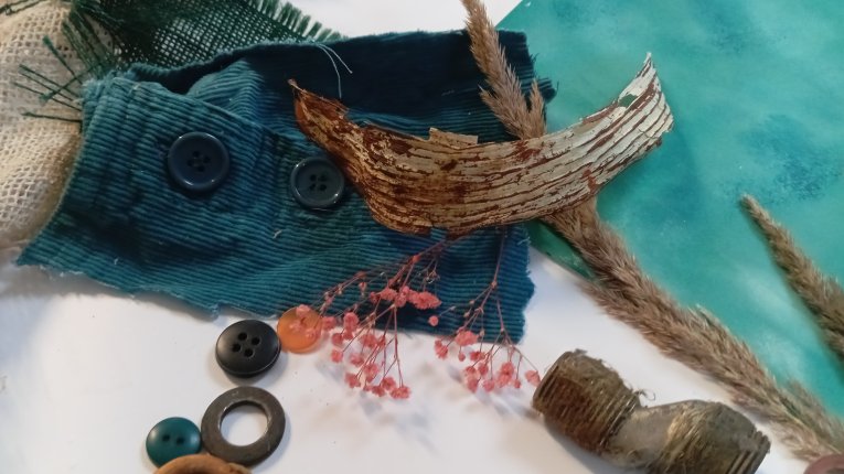 Rozsypane na białym tle materiały do tworzenia asamblażu. Widoczne są: fragmenty niebieskiego sztruksu z guzikami, kawałek zielonej siatki, suchy kłos zboża, kawałek zwiniętej kory brzozowej, drobne różowe kwiatuszki (gipsówka), metalowa złączka rury oraz