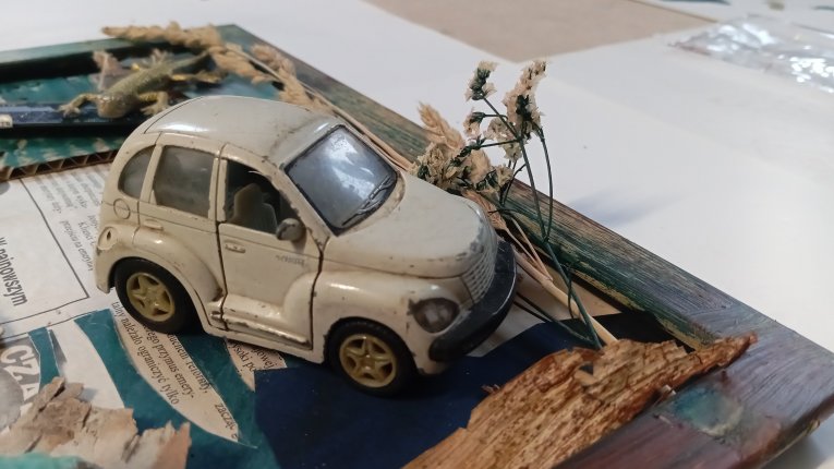 Zdjęcie wykonane pod kątem, skupiające się na detalu białego zabawkowego samochodu (model przypominający Chryslera PT Cruisera), który jest przytwierdzony do wnętrza ramy. Samochód jest zakurzony i nosi ślady użytkowania. Obok niego znajdują się suche kło
