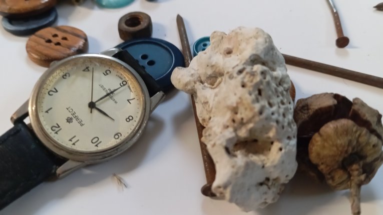 Zbliżenie na grupę przedmiotów: klasyczny zegarek naręczny marki „Perfect” na czarnym pasku, duży kawałek jasnego pumeksu lub porowatego kamienia, zasuszony owoc (orzech), kilka rdzawych gwoździ oraz różnokolorowe guziki porozrzucane na białym blacie.