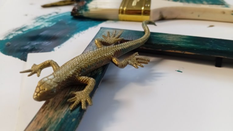 Zbliżenie na narożnik pomalowanej na zielono ramy, na której siedzi złota, plastikowa figurka jaszczurki. W tle widać białe podłoże z plamami morskiej farby, pędzle malarskie (jeden szeroki z jasnym włosiem) oraz słoiczek złotej farby. Zdjęcie wykonane z