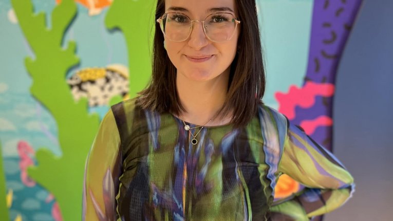 Osoba dorosła, kobieta, w kostiumie inspirowanym motywami podwodnymi, stojąca na tle kolorowej scenografii ekspozycji.