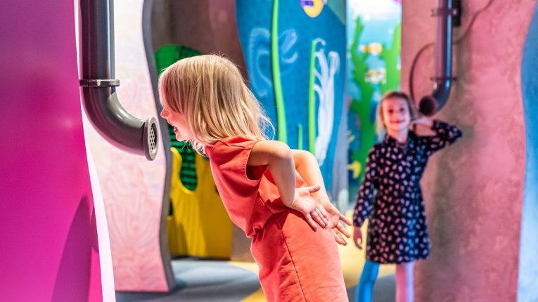 Dziecko pochylone przy instalacji edukacyjnej, mówiące do tuby dźwiękowej oraz drugie, słuchające dźwięków przez tubę umieszczoną w ścianie ekspozycji.