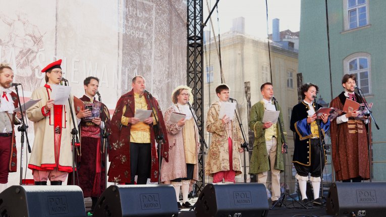 Grupa mężczyzn w historycznych strojach śpiewa na scenie plenerowej, stojąc przy mikrofonach i trzymając teksty w dłoniach.