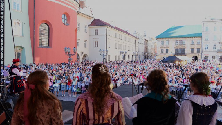 Widok ze sceny na licznie zgromadzoną publiczność na historycznym rynku. Na pierwszym planie stoją osoby w stylizowanych, historycznych strojach, śpiewające przed publicznością zgromadzoną na płycie rynku.