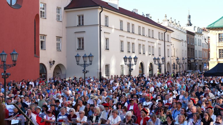 Widok na licznie zgromadzoną publiczność podczas wydarzenia na Małym Rynku w Krakowie. Tłum uczestników stoi przed sceną, część osób trzyma śpiewniki przygotowane na występ. W tle widoczna jest zabytkowa zabudowa rynku.